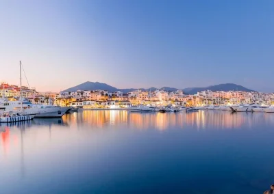 A tranquil view of Puerto Banús marina at dusk, featuring luxury yachts docked along the waterfront, white Mediterranean-style buildings illuminated with warm lights, and serene reflections on the calm water with mountains silhouetted in the distance.