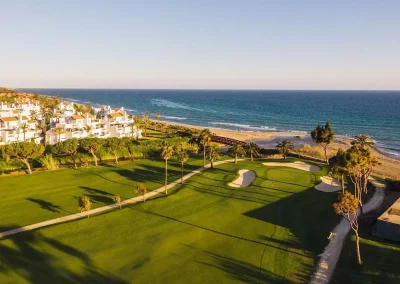A breathtaking view of Río Real's golf course featuring pristine fairways, sand bunkers, and towering palm trees. The course extends toward the coastline, where the sandy beach meets the ocean waves, creating a picturesque blend of green and blue under a bright sky.